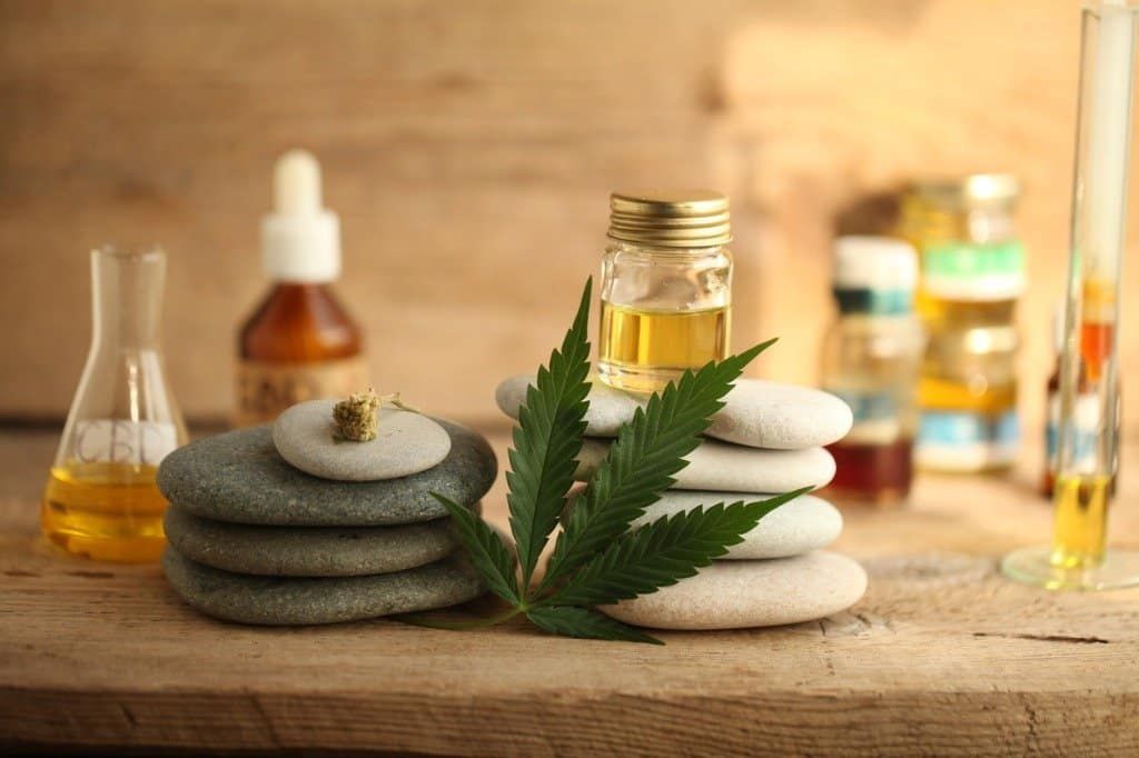 cannabis-leaf-resting-against-pile-of-stones-on-table-with-vials-and-bottles-of-cbd-and-other-essential-oils cannabis-leaf-resting-against-pile-of-stones-on-table-with-vials-and-bottles-of-cbd-and-other-essential-oils