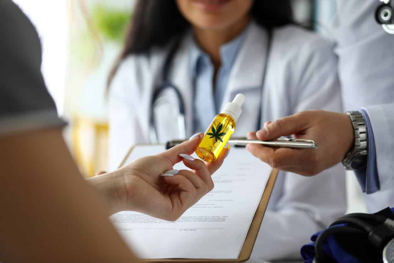 person-s-hand-holding-small-vial-of-cbd-oil-with-two-medical-professionals-in-background-wearing-white-coats-and-stethoscopes person-s-hand-holding-small-vial-of-cbd-oil-with-two-medical-professionals-in-background-wearing-white-coats-and-stethoscopes
